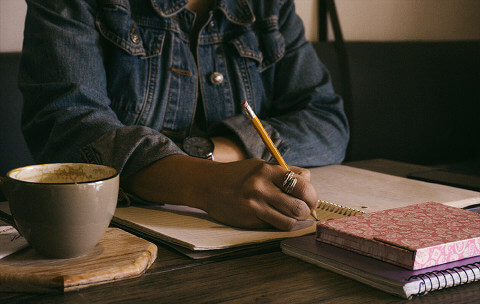 Person, sitting at a desk, making notes in a journal with a cup of coffee next to her