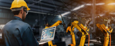 Man wearing overalls and hardhat, holding a laptop at an assembly line